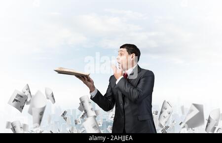 Embarrassé d'affaires couvrant la bouche avec la main et tenant un livre ouvert. L'homme d'affaires surpris en costume et cravate debout sur l'arrière-plan avec la chute de la ville Banque D'Images