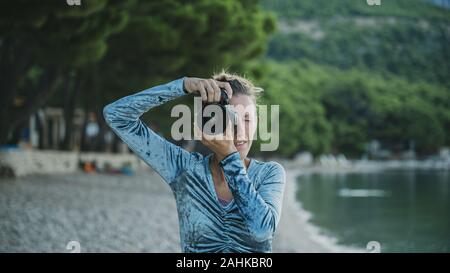 Vue de face d'une jeune femme prenant une photo directement à l'extérieur de l'appareil photo sur une plage de galets en début de matinée. Banque D'Images