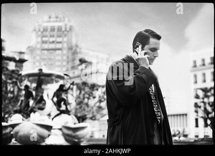 Vue latérale d'un homme parle sur son téléphone cellulaire avec une fontaine dans l'arrière-plan. Banque D'Images