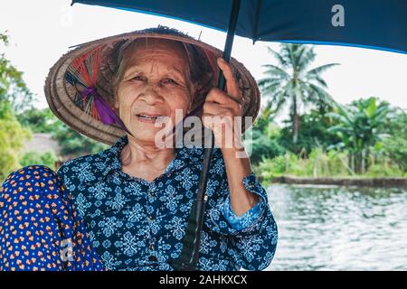 Une femme âgée utilise ses pieds pour pousser les rames d'une barque, tout en maintenant un ventilateur et parapluie, prenant les touristes sur la rivière Ngo Dong, Vietnam Banque D'Images