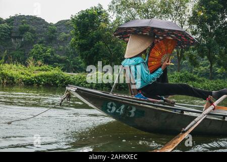 Une femme âgée utilise ses pieds pour pousser les rames d'une barque, tout en maintenant un ventilateur et parapluie, prenant les touristes sur la rivière à Hoa Lư, Vietnam Banque D'Images