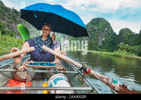 Une femme âgée utilise ses pieds pour pousser les rames d'une barque, tout en maintenant un ventilateur et parapluie, prenant les touristes sur la rivière Ngo Dong, Vietnam Banque D'Images