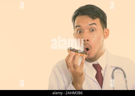 Studio shot of young Asian man eating donut médecin tout en regardant choqué Banque D'Images