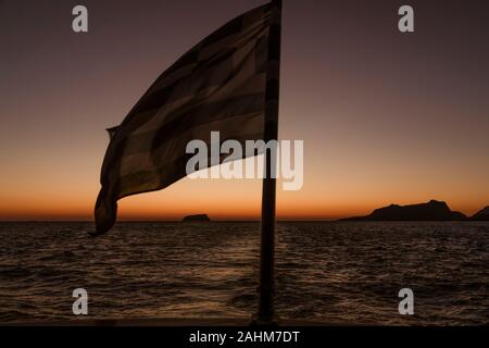 Le drapeau grec voltigeant dans le vent de l'arrière d'un bateau à voile avec un coucher de soleil en arrière-plan dans les îles grecques, près de l'île de Santorin en Grèce Banque D'Images