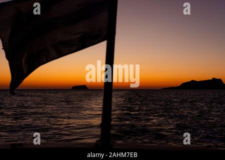 Le drapeau grec voltigeant dans le vent de l'arrière d'un bateau à voile avec un coucher de soleil en arrière-plan dans les îles grecques, près de l'île de Santorin en Grèce Banque D'Images