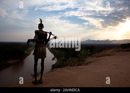 Guerrier de la tribu Karo peint sur tout le corps, avec de la fourrure sur la tête et avec une Kalachnikov, debout sur un plateau au-dessus de la vallée de la rivière Omo, Ethiopie, Banque D'Images