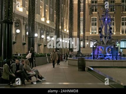 Des couples s'asseoir sur des bancs et des gens à pied le soir à Hays Galleria, le temps de Noël avec la sculpture de navigateurs en arrière-plan. Banque D'Images