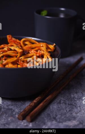 Les nouilles Udon avec de la viande de poulet dans un bol avec des baguettes et une tasse de thé sur fond de pierre sombre Banque D'Images