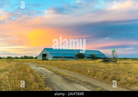 Route de campagne et l'ancien hangar de tonte en tôle ondulée sur le coucher du soleil près de Longreach, Queensland. Banque D'Images