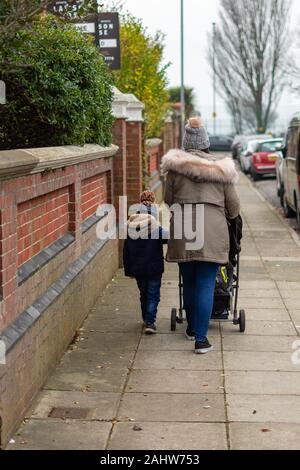 Une mère poussant un landau, une poussette ou une poussette dans la rue tandis qu'un jeune enfant marche aux côtés de holding sur Banque D'Images