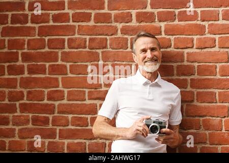 Smiling young man looking at camera. Avec appareil photo touristique debout à mur de briques. Banque D'Images