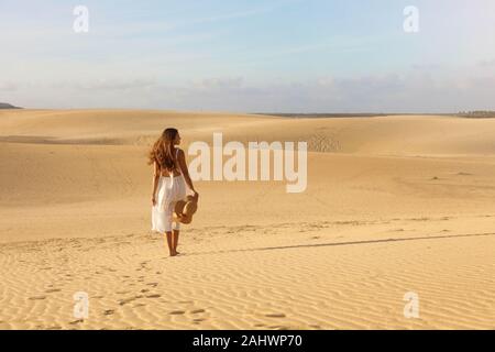 Belle jeune femme avec robe blanche marche dans les dunes du désert au coucher du soleil. Fille qui marche sur le sable doré sur Corralejo Beach, à Fuerteventura. Banque D'Images