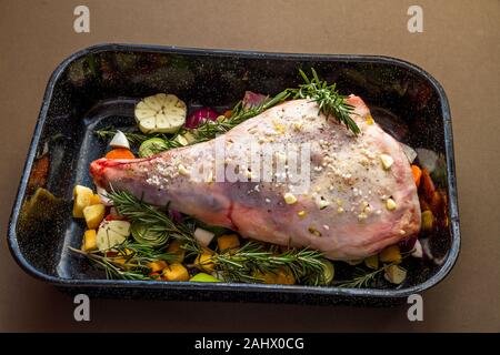 Le gigot d'agneau prêt à rôtir dans le bac avec du sel, l'ail, le romarin, les légumes et l'ail. Banque D'Images