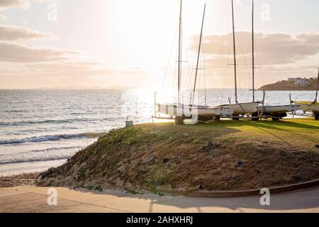 Bateaux à voile garé sur l'herbe à côté du club-house, tôt le matin avec la lumière qui se reflète sur l'océan Banque D'Images