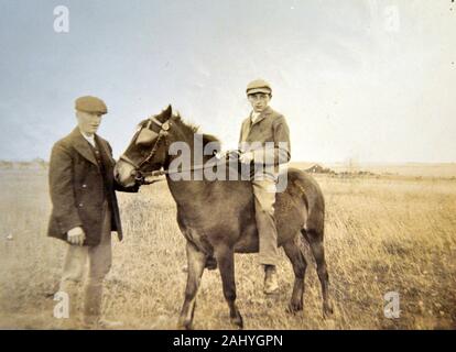 Photographie sépia ancienne, antique, noire et blanche d'un garçon assis sur un cheval qui est tenu par un homme dans un champ à la campagne Banque D'Images