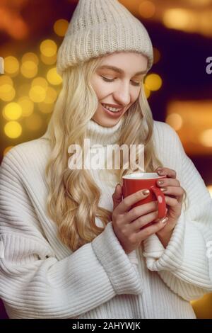 Jeune femme bénéficiant d'une tasse de café chaud en hiver il saigner dans ses mains en verre, avec un sourire béat contre un orange chaud backgroun Banque D'Images