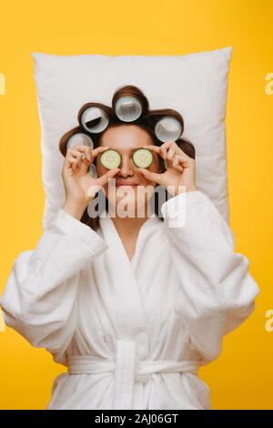 Femme se trouve sur l'oreiller avec cheveux en rouleaux holding tranches de concombre sur les yeux Banque D'Images