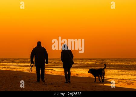 Couple de propriétaires de chien à marcher le long de la côte avec chien sans sur une plage de sable, silhouetté contre le ciel orange au coucher du soleil sur une soirée froide en hiver Banque D'Images