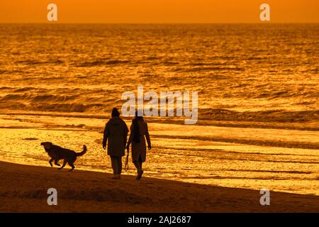 Deux femmes marchant avec chien sans le long de la côte sur une plage de sable au coucher du soleil sur une soirée froide en hiver Banque D'Images