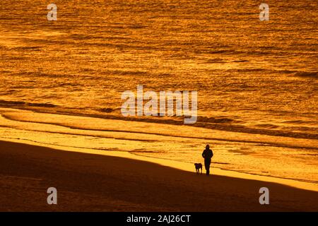 Propriétaire de chien solitaire marchant le long de la côte avec chien sans sur une plage de sable, en silhouette au coucher du soleil sur une soirée froide en hiver Banque D'Images