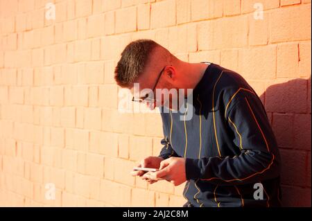 Guy smiling positive dans casualwear avec téléphone mobile. Young caucasian man standing at outdoor mur rouge et voir des vidéos drôles dans son appareil. Loisirs, fu Banque D'Images