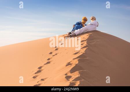 Portrait d'un Bédouin nomad avec turban coloré et grand sourire assis sur dune de sable touristique populaire Merzouga désert. Homme Touareg Banque D'Images