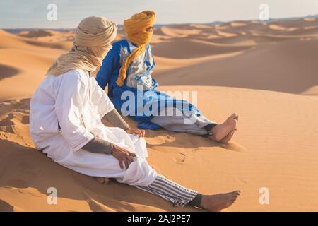 Portrait d'un Bédouin nomad avec turban coloré et grand sourire assis sur dune de sable touristique populaire Merzouga désert. Homme Touareg Banque D'Images
