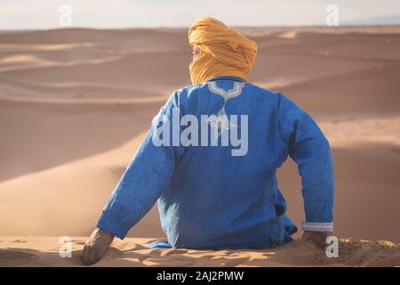 Portrait d'un Bédouin nomad avec turban coloré et grand sourire assis sur dune de sable touristique populaire Merzouga désert. Homme Touareg Banque D'Images