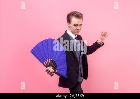 Portrait du jeune magicien concentré grave in tuxedo standing avec bras soulevé et holding blue ventilateur, illusioniste montrant tour de magie, de performance. Banque D'Images