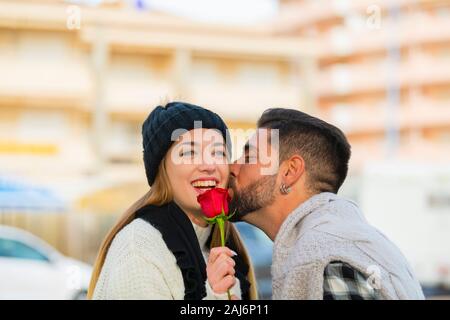 Jeune femme tenant une rose rouge rit comme elle reçoit un baiser romantique sur la joue par un jeune homme sur un arrière-plan flou. Concept de la Saint-Valentin. Banque D'Images