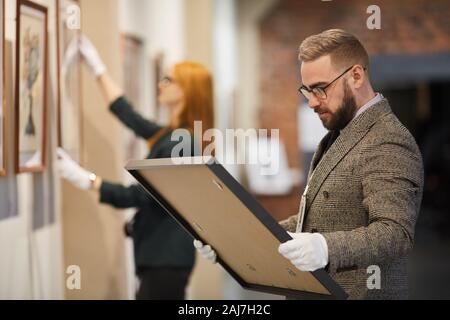 Gestionnaire en fonction de l'art et dans les gants d'un permanent à la recherche de photo dans ses mains alors que woman hanging peinture sur le mur de la galerie Banque D'Images
