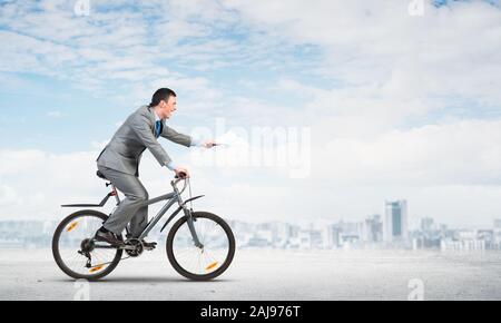 Businessman with documents papier dans la main en vélo. Date limite pour la paperasse. Personnel de l'entreprise en costume gris vélo sur fond de ciel Banque D'Images