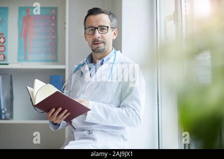 Portrait of mature homme médecin en blouse blanche assis avec notes et contrôler ses notes à l'office de tourisme Banque D'Images