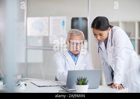 Médecin Senior en robe blanche assise à la table fonctionne sur les ordinateurs portables et de discuter avec son infirmière at office Banque D'Images