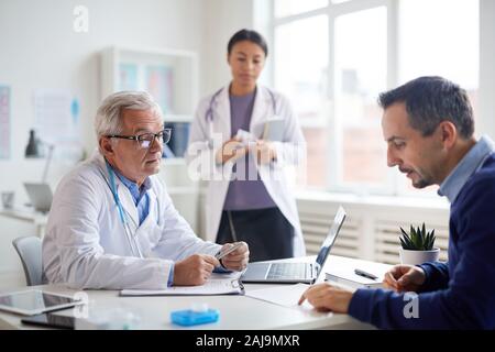 Hauts homme médecin en blouse blanche à parler au patient à la table qu'ils discuter de méthode de traitement à l'infirmière dans l'arrière-plan Banque D'Images