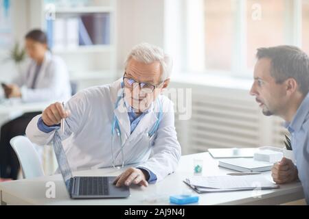 Médecin à lunettes assis à la table de pointage et de discuter de la maladie avec son patient Banque D'Images