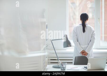 Vue arrière du jeune femme médecin en blouse blanche debout et regardant par la fenêtre à l'office de tourisme Banque D'Images