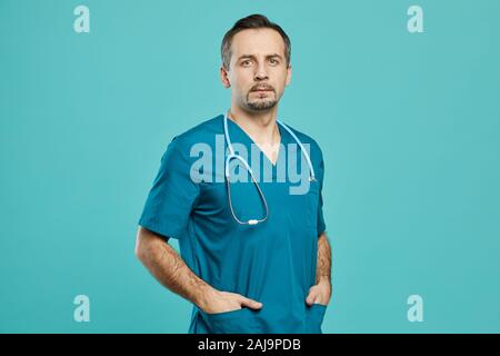 Portrait of mature male doctor in scrubs et stéthoscope standing dans la poche et à la caméra à l'encontre de l'arrière-plan bleu Banque D'Images
