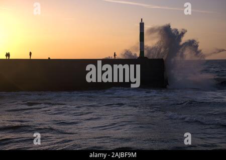 Brisant les vagues contre la pierre jetée à Aberystwyth, avec des gens debout sur la jetée et le coucher de soleil. Banque D'Images