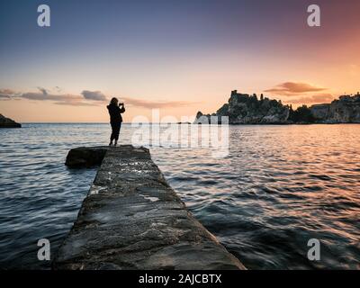 Silhouette d'une jeune fille photographiant la côte sicilienne au coucher du soleil, Isola Bella, Taormina, Italie Banque D'Images