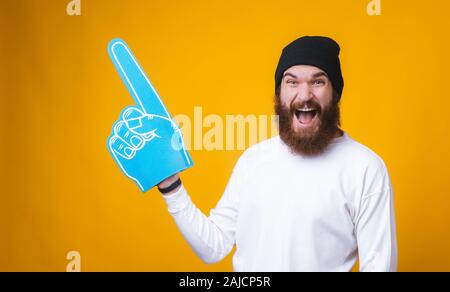 Jeune homme barbu est dans l'étonnement et la tenue d'un grand ventilateur gant et dirigés à l'écart près de mur jaune. Banque D'Images