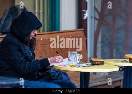 Genève, Suisse - le 14 avril 2019 : un homme avec une capuche sur sa tête, est assis à une table dans un café en plein air, le tabagisme et la rédaction d'un document - droit Banque D'Images