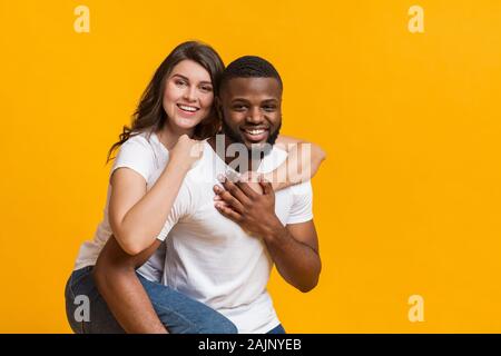 Guy Afro piggyback ride donnant à sa petite amie, jeune couple interracial avoir du plaisir ensemble, posant sur fond jaune avec de l'espace libre Banque D'Images