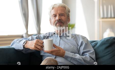 Elderly man sitting on sofa holding cup looking at camera Banque D'Images