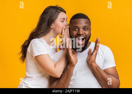 Jeune femme secret partage avec son petit ami noir choqué, commérer ensemble, posant sur fond jaune Banque D'Images