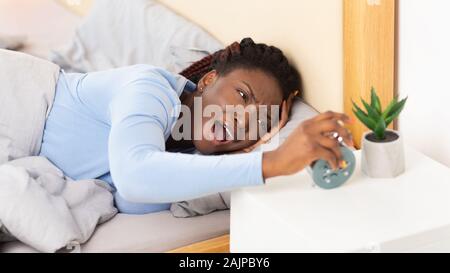 Mauvais matins. Fille noire La mise hors tension de Alarm-Clock La tête touchant un moment difficile le Réveil au lit à l'intérieur. Panorama Banque D'Images