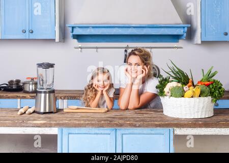 Jeune mère et fille joyeuse peu penchée sur la table, debout dans la cuisine avec des meubles modernes, panier de légumes frais sur la table, l'éco-ami Banque D'Images