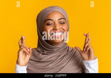 Veuillez, s'il vous plaît, s'il vous plaît. Closeup portrait of dreamy black jeune musulmane en hijab crossing fingers, rendant le souhaitent, sur fond jaune Banque D'Images