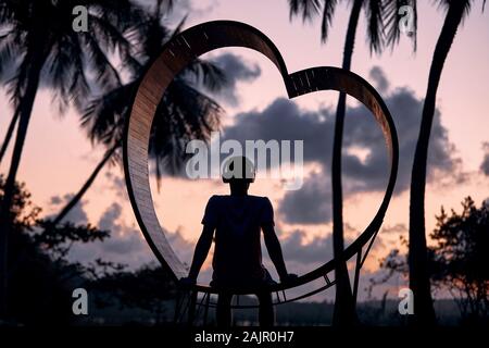 Détente dans un paradis tropical. Jeune homme assis avec un casque en bois en plein coeur et l'écoute la musique contre palmiers au beau coucher du soleil. Banque D'Images