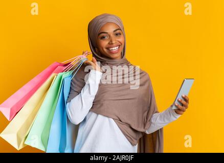 Magasinage en ligne App Concept. Portrait de jeune fille en noir joyeux hijab islamique avec le smartphone et sacs lumineux sur fond jaune Banque D'Images